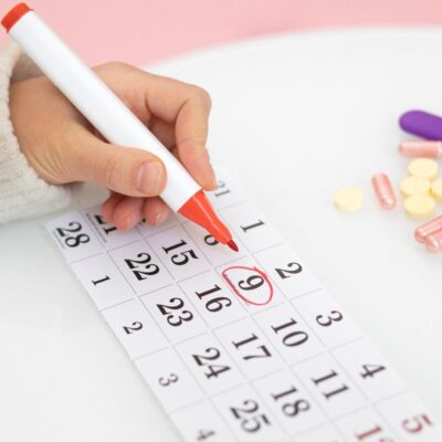 Kobieca dłoń zaznaczająca datę w kalendarzu leżącym na blacie z rozsypanymi tabletkami.