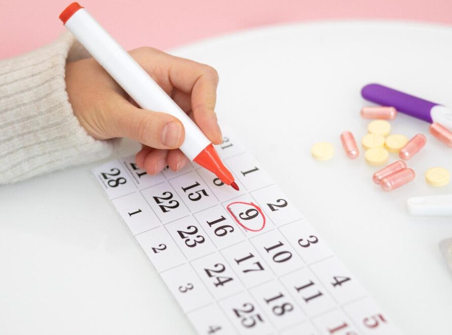 Kobieca dłoń zaznaczająca datę w kalendarzu leżącym na blacie z rozsypanymi tabletkami.